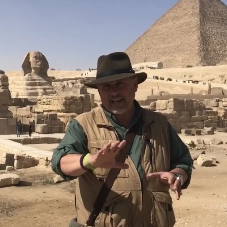 A man in a hat and vest standing next to the pyramids.