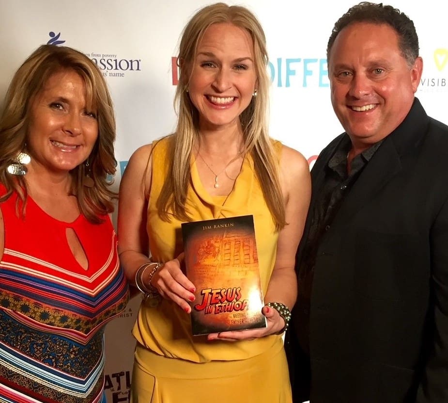 A woman holding an orange book with two men standing next to her.