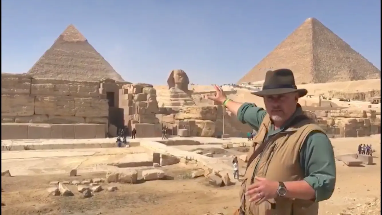 A man in a hat standing next to the pyramids.