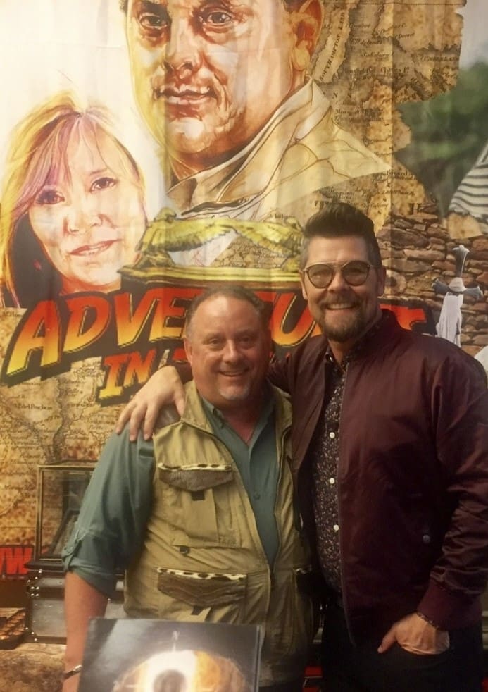 Two men standing in front of a poster for adventure in the woods.